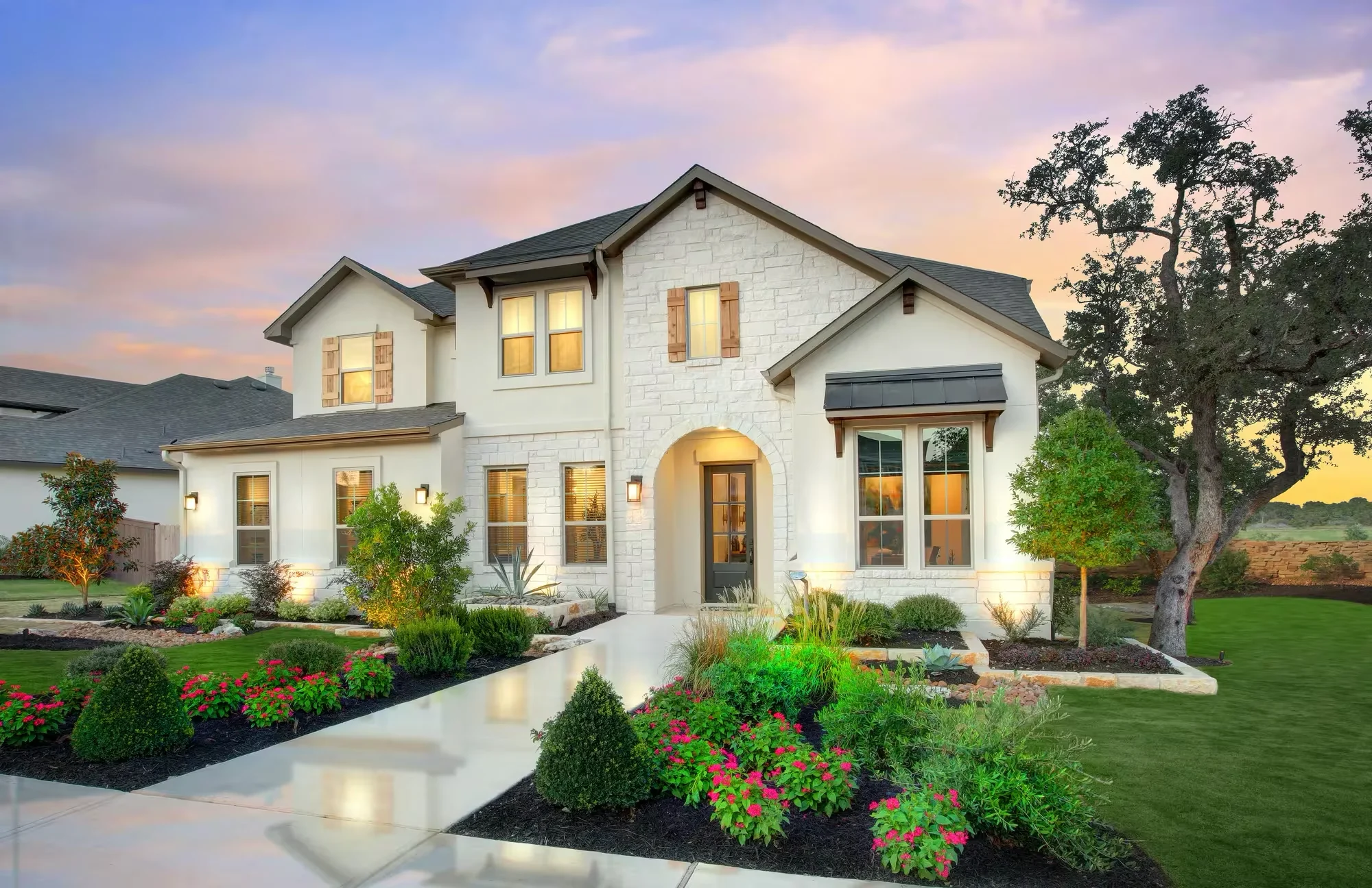 Exterior of a two-story Texas Hill Country home in Provence with white stone, wood shutters, landscaped flower beds, and a spacious green lawn.